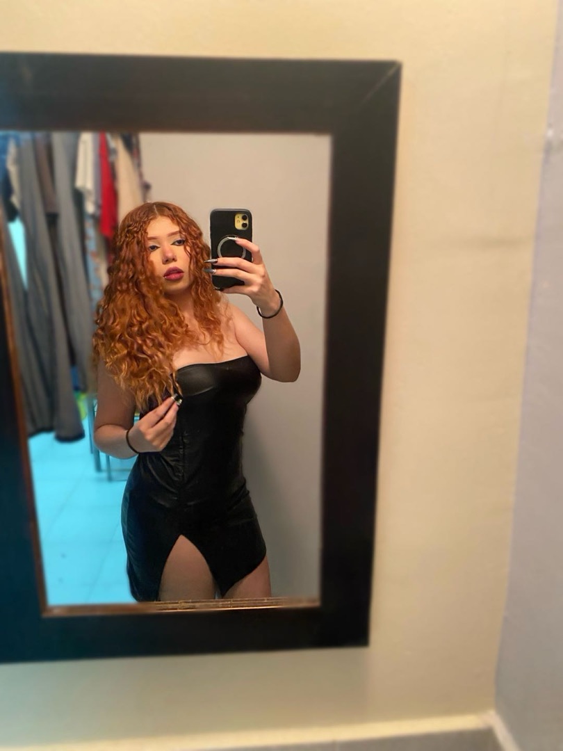 Woman with curly red hair taking a mirror selfie in a black dress